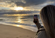 © Angelov - Woman holding mobile phone in hand and taking sunrise photo. Woman using smart phone take a photo at evening on the beach of sunset. Travel photo, copy space.