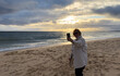 © Angelov - Woman holding mobile phone in hand and taking sunrise photo. Woman using smart phone take a photo at evening on the beach of sunset. Travel photo, copy space.