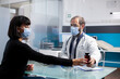 © DC Studio - Young woman sits in clinic appointment as doctor explains painkillers and healthcare supplement. Physician giving prescription pills bottle to female patient through protective screen on table.