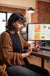 © DC Studio - Businesswoman checking emails on smartphone as desktop monitor displays business analytics at office desk. Female employee holding mobile device chatting with family and friends in brick wall room.