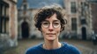 © Newleks - Young woman with short curly hair, round glasses and freckles stands on a cobblestone city street, looking at camera with a confident, thoughtful, authentic expression