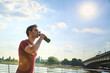 © Teodor Lazarev - Young athletic man drinking water outdoors, staying hydrated and refreshed during a hot summer day, with a river and bridge visible in the urban landscape