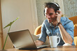 © Teodor Lazarev - Smiling man wearing headphones and using a laptop for remote work or online communication at home, enjoying music or a video call over his morning coffee