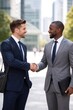 © Татьяна Евдокимова - Two diverse businessmen smiling and shaking hands outdoors in a modern urban setting, sealing a successful agreement that highlights partnership, trust, and professional collaboration