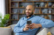 © Liubomir - Smiling man relaxing at home on a comfortable sofa, holding a smartphone and looking away, enjoying leisure time and digital connection in a modern living room