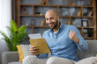 © Liubomir - Excited man at home celebrates good news after opening a mail letter, sitting on a sofa with fist raised, smiling in relief and joy over an approval or success notification