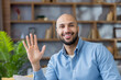 © Liubomir - Young bald man with beard wearing a blue shirt, smiling at the camera and waving his hand in a friendly gesture, sitting indoors in a modern office or living room setting