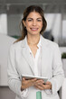 © fizkes - Vertical portrait young Hispanic businesswoman holding tablet looking at camera with smile enjoying using modern digital tools corporate business software in professional office work with confidence