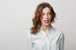 © SHOTPRIME STUDIO - Young woman with curly hair wearing white shirt makes funny face sticking out tongue looking sideways. Studio portrait on light background with casual style and playful mood concept.