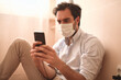 © Teodor Lazarev - Young man in a medical face mask sits at home, focused on his smartphone, connecting with others during quarantine and practicing social distancing for safety and health