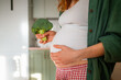 © wifesun - Expecting woman embracing her pregnancy, holding a fresh broccoli floret, symbolizing healthy eating habits and incorporating a nutritious diet for prenatal wellness and baby development