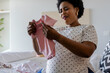 © wavebreak3 - African American pregnant woman sitting on bed holding pink onesie, inspecting baby garments