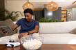 © wavebreak3 - African American adult man sitting on sofa holding tablet and snacking from popcorn, gamepad nearby