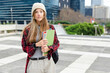 © wavebreak3 - Woman standing holding notebooks and folders on urban plaza wearing beanie, backpack, smartwatch