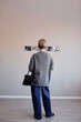 © Seventyfour - Vertical shot of middle aged Caucasian woman standing with back to camera observing black and white photographs on wall in gallery setting, holding handbag in left hand, short hair visible