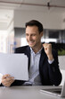 © fizkes - Hispanic corporate executive checking business papers at office workplace excited about successful sales agreement celebrate deal approval lucrative contract terms conditions triumph. Vertical shot