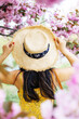 © Mykola - A high-angle, rear-view photograph of a woman wearing a wide-brimmed straw hat and a yellow floral dress, looking into a vibrant canopy of pink cherry blossoms. The woman’s hands are gracefully holdin