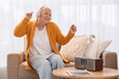 © Pixel-Shot - Senior woman in headphones listening to radio on sofa at home