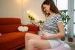 © wifesun - Pregnant woman engaging in a prenatal workout at home, using a pink fitness ball while looking at her smartphone for exercises, embracing health and wellness during expectant motherhood