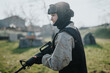 © qunica.com - Female airsoft player stands in tactical gear with protective goggles. She holds a rifle ready in an open field under daylight.