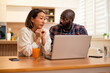 © djoronimo - Adult couple sits together at a wooden table, using laptop for video communication in a bright and cozy home kitchen.