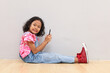 © ArvStd - Portrait of a 7-8 year old girl sitting on the floor holding and using a mobile phone, smiling looking at the camera, isolated on a grey background.