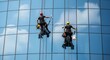 © Iqbal - Two professional high-rise window cleaners suspended by ropes, meticulously cleaning the reflective glass facade of a modern office building on a clear sunny day.