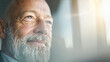 © Kowit - A close-up of an elderly man with a white beard gazes thoughtfully out of a window with soft sunlight illuminating his face.