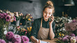 © Kowit - A smiling florist wearing an apron talks on the phone while writing notes, surrounded by colorful flowers in a cozy flower shop.
