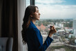 © Parichat - Young Asian woman holding a coffee cup, looking thoughtfully at the city view from a hotel window