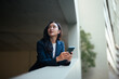 © Parichat - Young businesswoman holding smartphone looking up thinking future