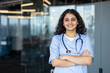 © Liubomir - Young female medical professional in scrubs and stethoscope smiling, standing with arms crossed at a modern clinic or hospital, representing healthcare, wellness, and dedicated service