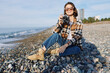 © SHOTPRIME STUDIO - Female photographer on a pebble beach with a camera, casual plaid jacket and boots, sunny day, sea in the background, capturing moments and enjoying outdoor photography.