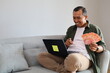 © ArvStd - Adult man sitting on sofa at home holding Indonesian banknotes, staring and pointing thoughtfully at laptop screen while deciding on expenses, considering purchases, investments or financial planning.