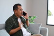 © ArvStd - An adult man is talking on his cell phone while sitting on the sofa at home with a shocked expression while holding his chest.