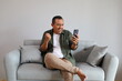 © ArvStd - Adult man sitting on sofa at home happy using smartphone, celebrating success, victory, winning lottery jackpot, goal achievement, good and positive news, while raising clenched fists.