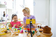 © sofiko14 - A mother and daughter wearing bunny ears paint Easter eggs together at a kitchen table while recording with a smartphone on a tripod