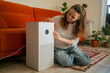 © wifesun - Woman sitting on the floor adjusting the display settings on a smart air purifier, bringing fresh air and better breathing to her modern home environment, prioritizing health technology and comfort