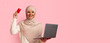 © Prostock-studio - Woman with a headscarf is smiling while raising a credit card in one hand and holding a laptop in the other. She is in a simple area with a pink background, focused on her task.