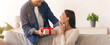 © Prostock-studio - A man hands a wrapped gift to a woman while they sit on a couch in a living room. The woman smiles and looks surprised as the man presents the gift. Green plants are in the background.