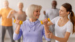 © Prostock-studio - A group of older adults participates in a strength training class led by a trainer. One woman lifts yellow weights while the trainer assists her. The atmosphere is supportive and energetic.