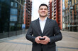© muse studio - Confident young man in a business suit stands smiling, holding a smartphone on a modern urban walkway
