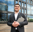 © muse studio - Confident young businessman in a dark suit holding a tablet standing before a modern glass building on a bright day