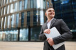 © muse studio - Young professional smiles holding laptop outside modern office building in a business district