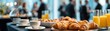 © Olga Ко - Ultra-wide banner of a corporate breakfast catering table with croissants, coffee, and juice. Blurred business people networking at a conference coffee break in the background.
