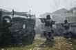© qunica.com - Paintball players in tactical vests and masks fire markers on a barrier wall of stacked tires. An outdoor paintball game scene shows teammates advancing under cover in a dusty field.