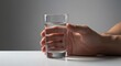 © Setyawan - A hand holds a clear glass of water on a white surface against a neutral backdrop