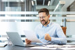 © Liubomir - Businessman in modern office celebrates good news, clutching a document and clenching his fist with a joyful, triumphant smile at his desk, laptop nearby, dressed in professional attire