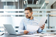 © Liubomir - Professional man wearing glasses sitting at a desk in a contemporary office, carefully reviewing multiple business documents and paperwork next to a laptop, focusing intently on analysis and data