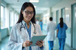 © Raisa - Asian female doctor uses tablet in hospital hallway. Medical pro smiles while looking at device. Healthcare worker with stethoscope checks digital info. Colleagues walk past in clinic.
